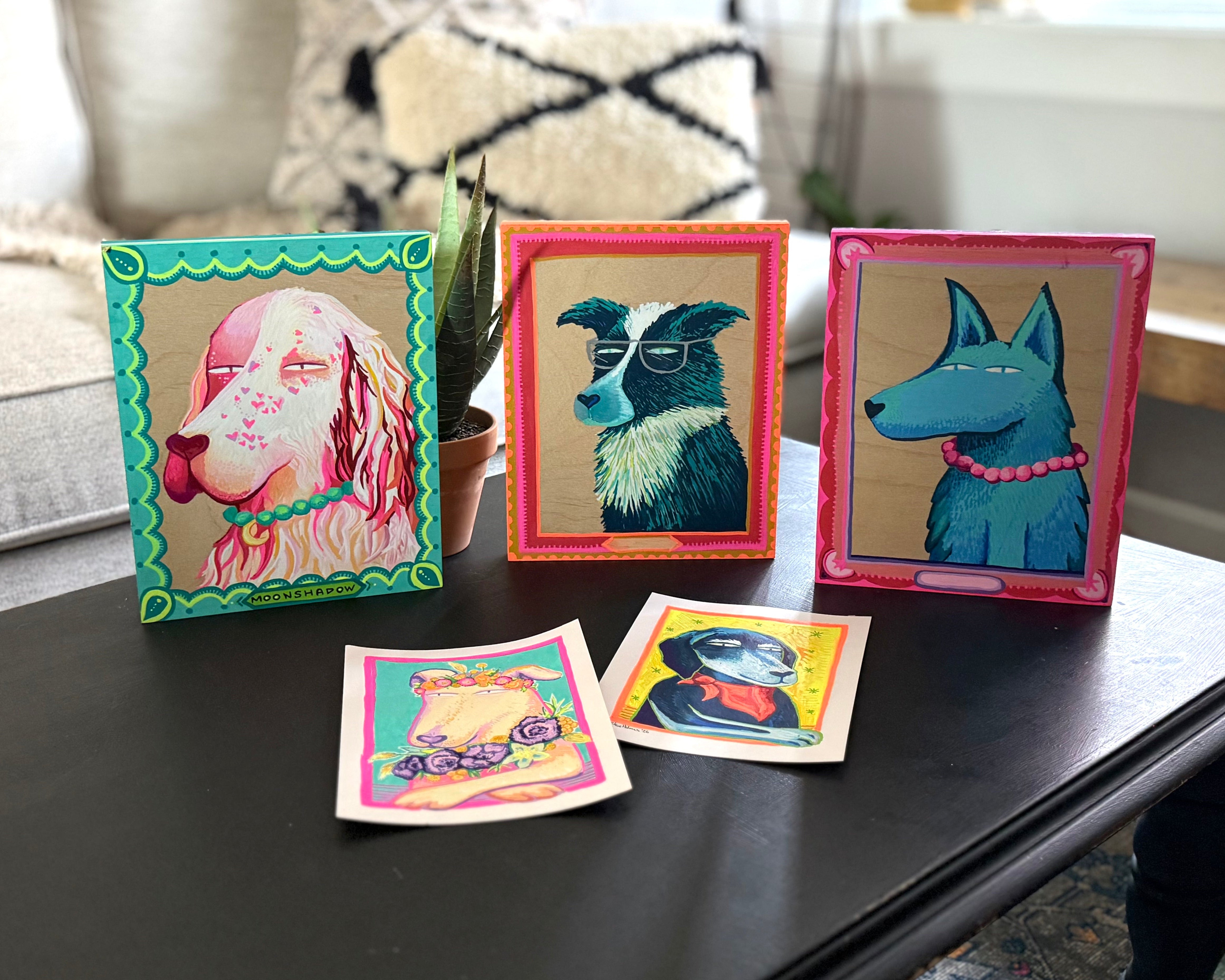 Three colorful framed art prints of stylized dogs displayed on a dark coffee table in a living room setting, with two small matching art cards in front.