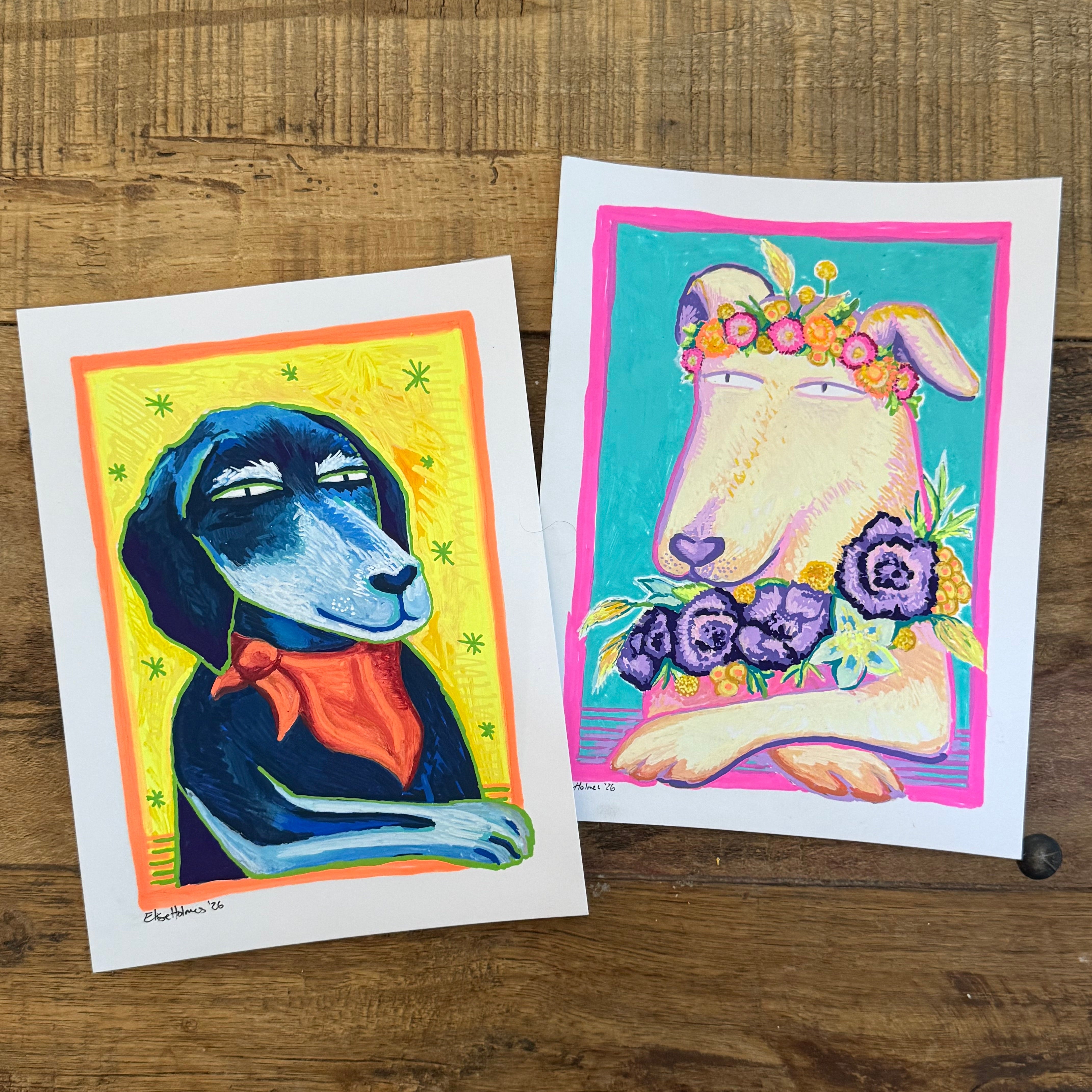 Two colorful art prints featuring stylized dog portraits, displayed on a rustic wooden tabletop. The left print shows a black dog with a red bandana on a yellow background, and the right print shows a cream-colored dog with purple flowers and a floral crown on a turquoise background.