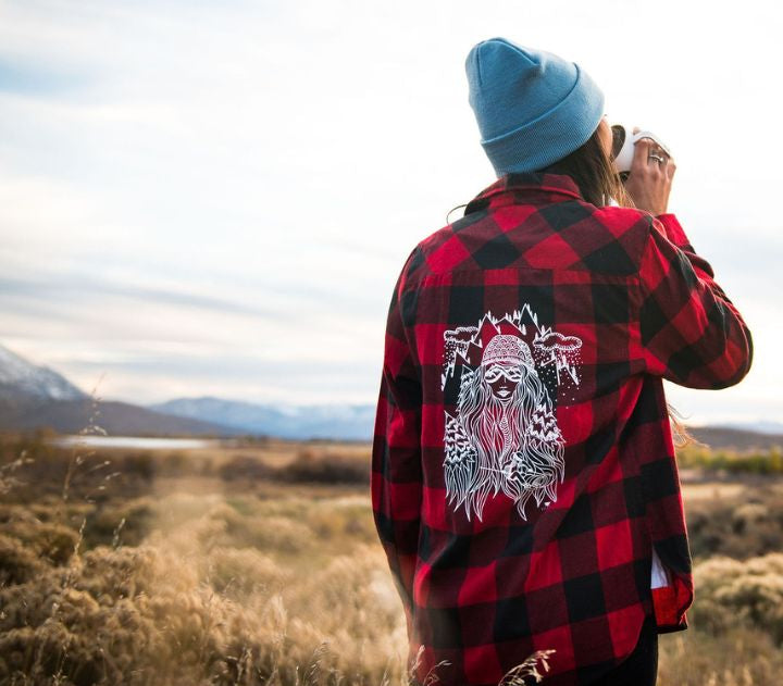 Person wearing a red and black checkered shirt with a graphic design, standing in a field with mountains in the background.