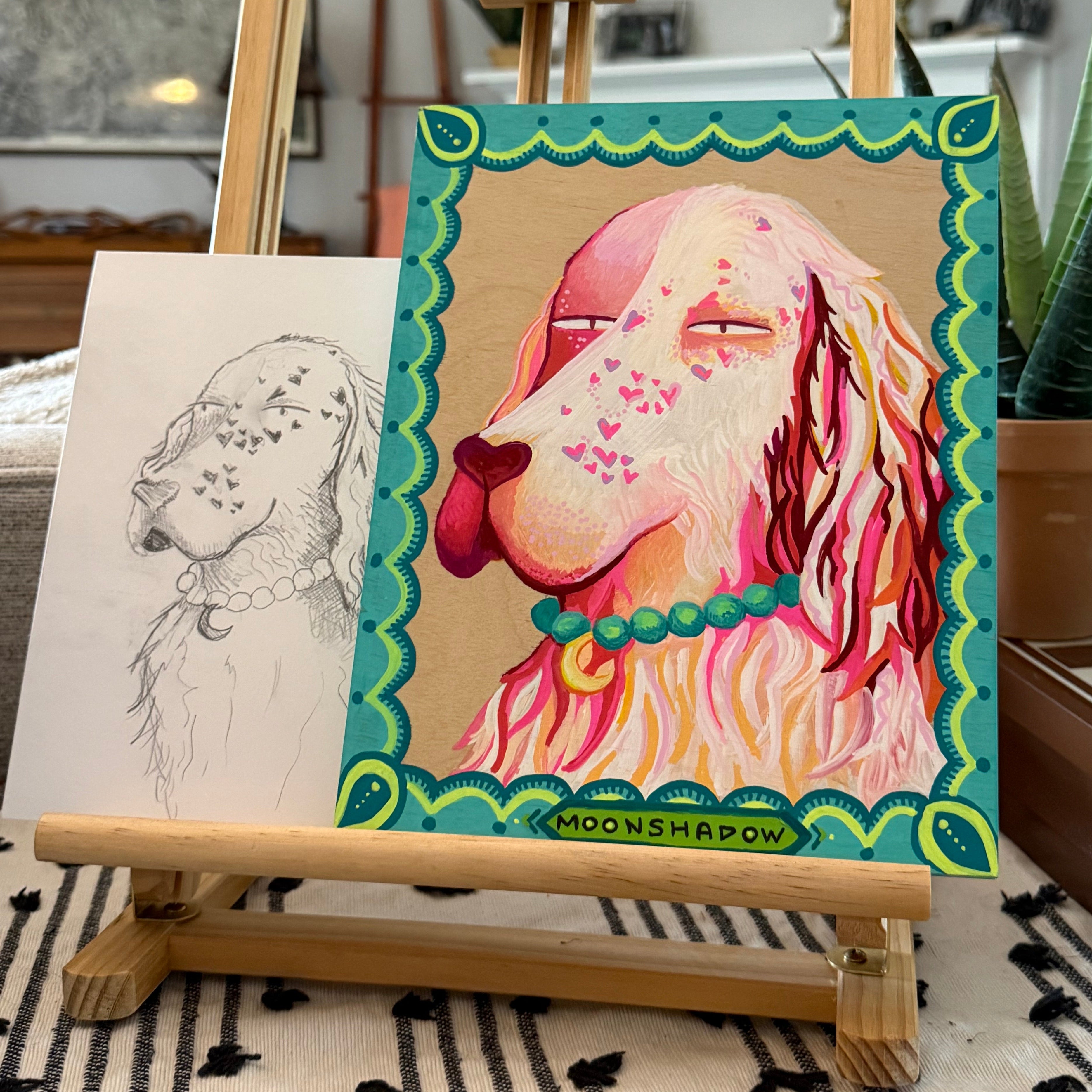 A colorful art print featuring a stylized illustration of a dog's face, displayed on a small wooden easel in an indoor setting next to a patterned rug and potted plant.