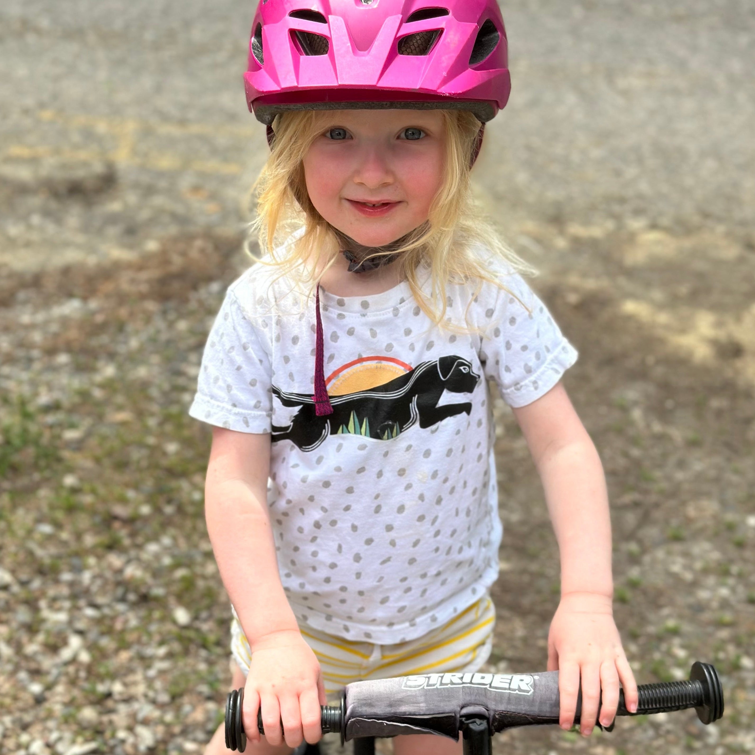 Child wearing a pink helmet and white shirt with a black animal design, standing on a gravel surface.